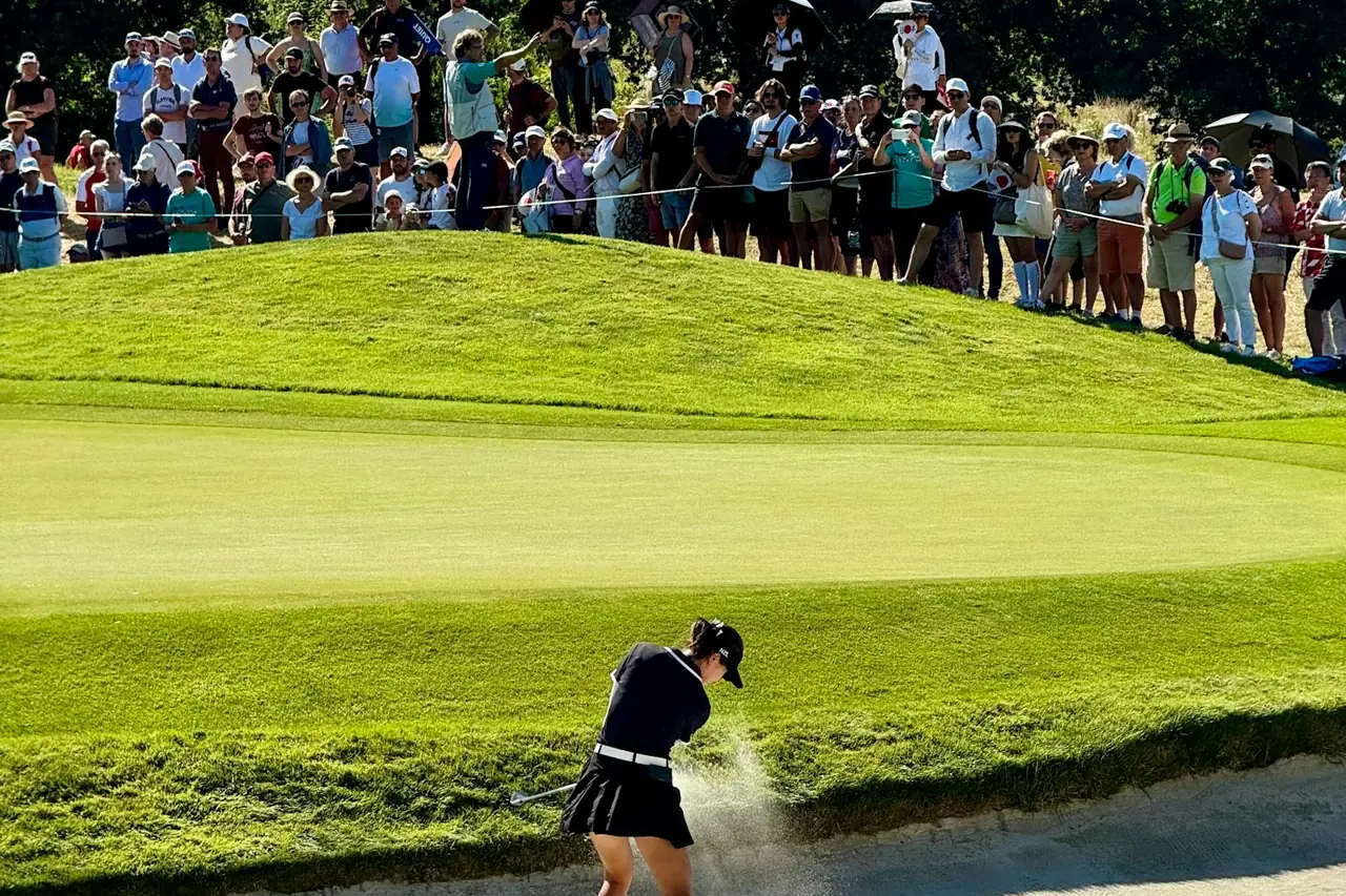A golfer plays out of a bunker in front of a tournament crowd